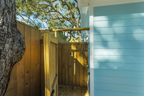 A wooden outdoor shower area with a tree trunk on the left, a wooden fence, and a blue building in the background. - Porch Light Cottage - Historic District Walk to the Beach, Private Pool, Spa, Fire Pit & Guest House (Saint Simons Island)
