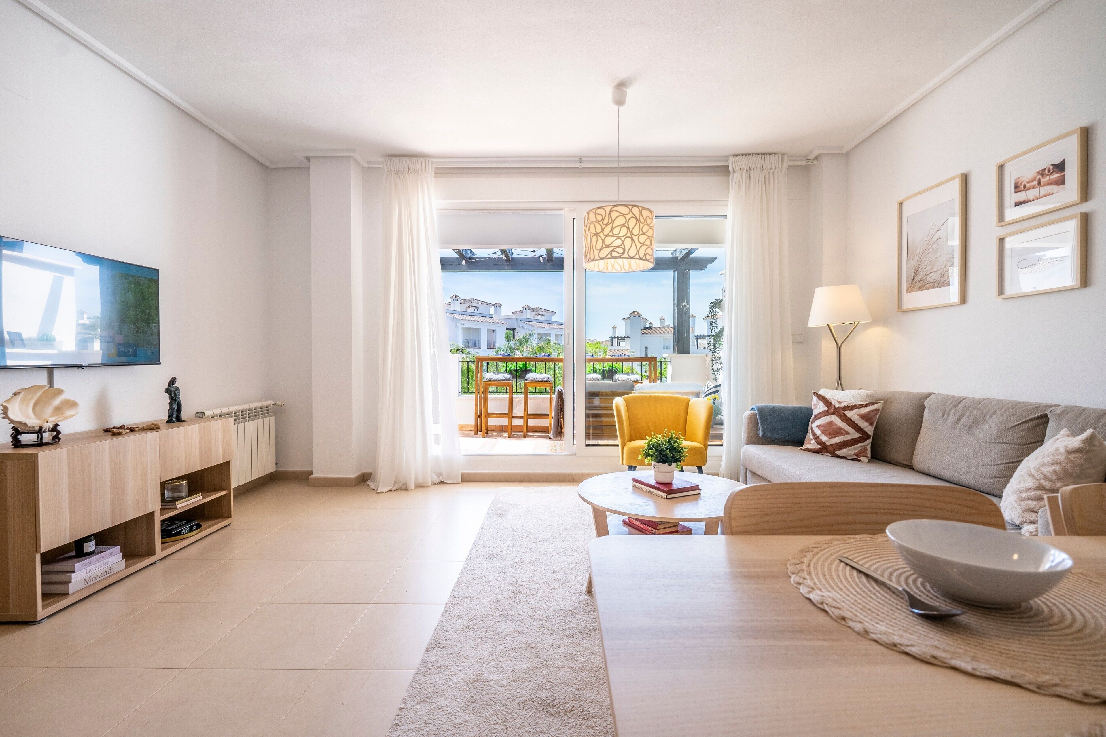 A modern living room with a sofa, coffee table, and TV. There's a balcony with a view, a dining area, and a lamp.