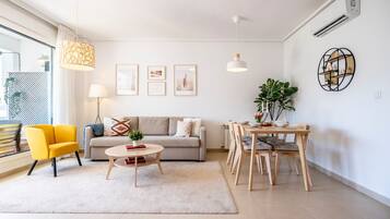 Een moderne woonkamer met een bank, gele fauteuil, rond salontafeltje, eettafel met stoelen, hanglampen, ingelijste foto's en een potplant.