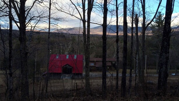 Ein rotes Bauernhaus in einer bewaldeten Landschaft mit Bergen im Hintergrund.