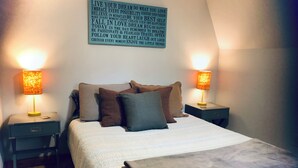 Ein ordentlich bezogenes Bett mit einer Tagesdecke und Kissen, zwei Nachttische mit Lampen und ein motivierendes Zitat an der Wand.