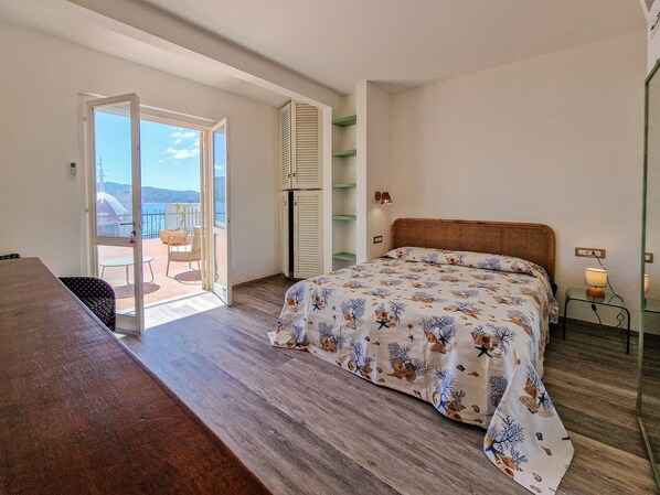 A bedroom with a bed, a wooden desk, a chair, a balcony with a view, and a shelf with decorative items.