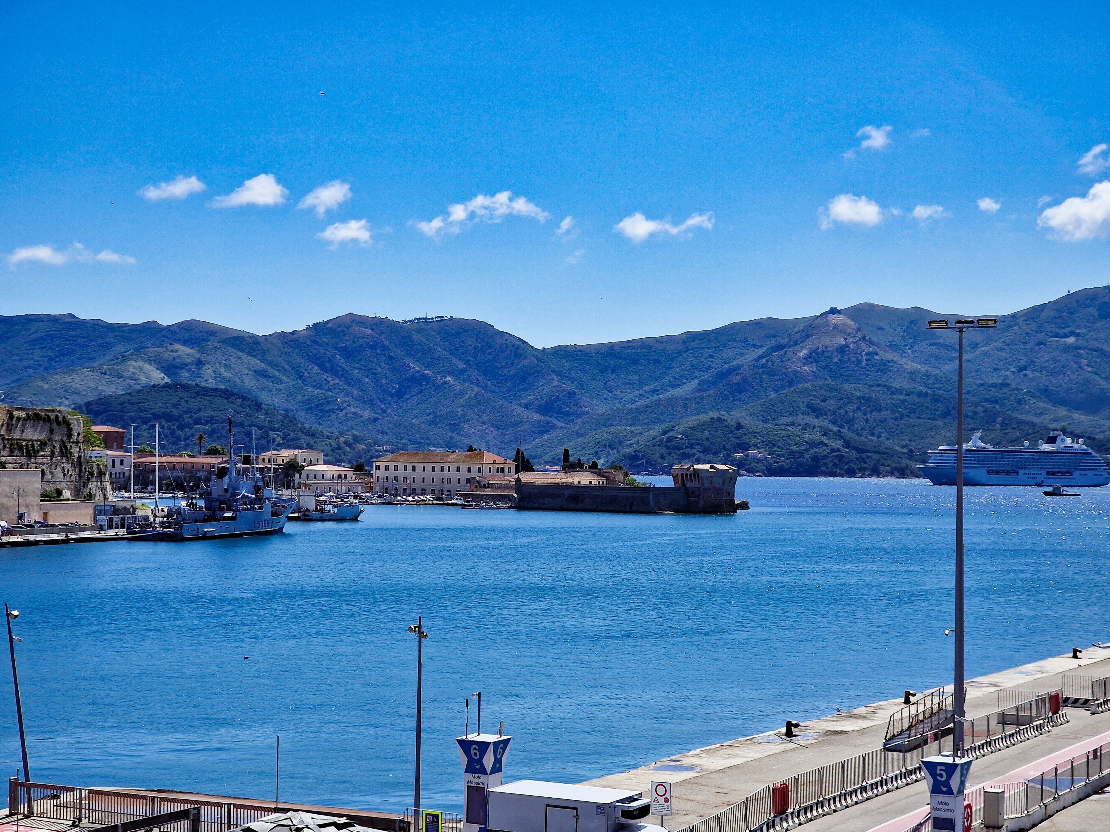 A coastal marina with boats, a lighthouse, and a mountainous backdrop.