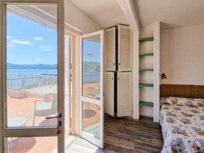 A room with a bed, a balcony with a view of the sea, and a sliding door leading to an outdoor area.