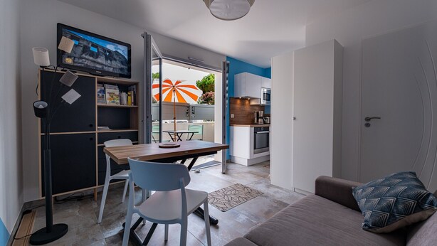 Una sala de estar moderna con un sofá, una mesa de comedor con sillas, un televisor montado en la pared y una cocina visible a través de una puerta abierta.
