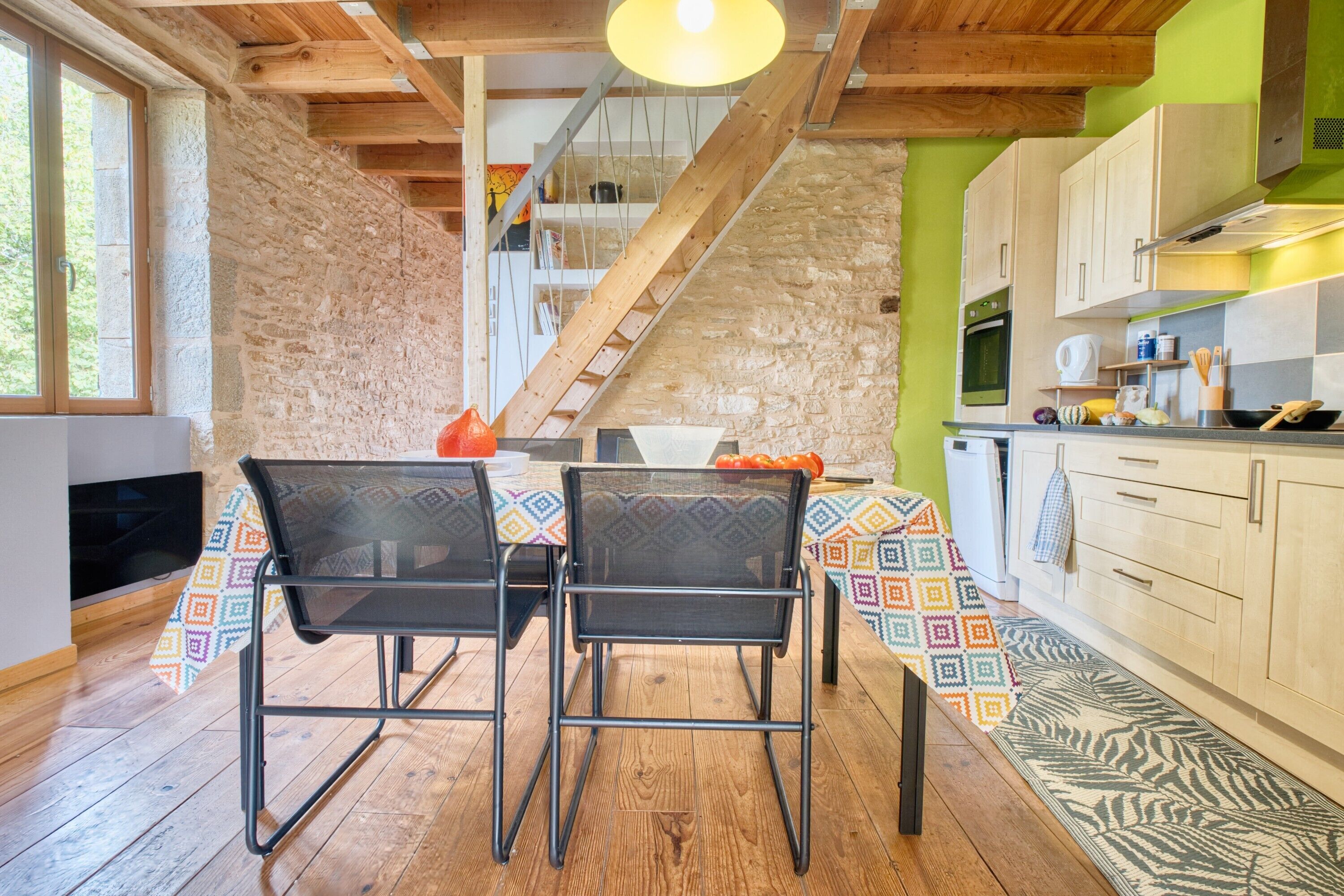 A rustic kitchen with a wooden staircase, a dining table with chairs, and a view of the outdoors.