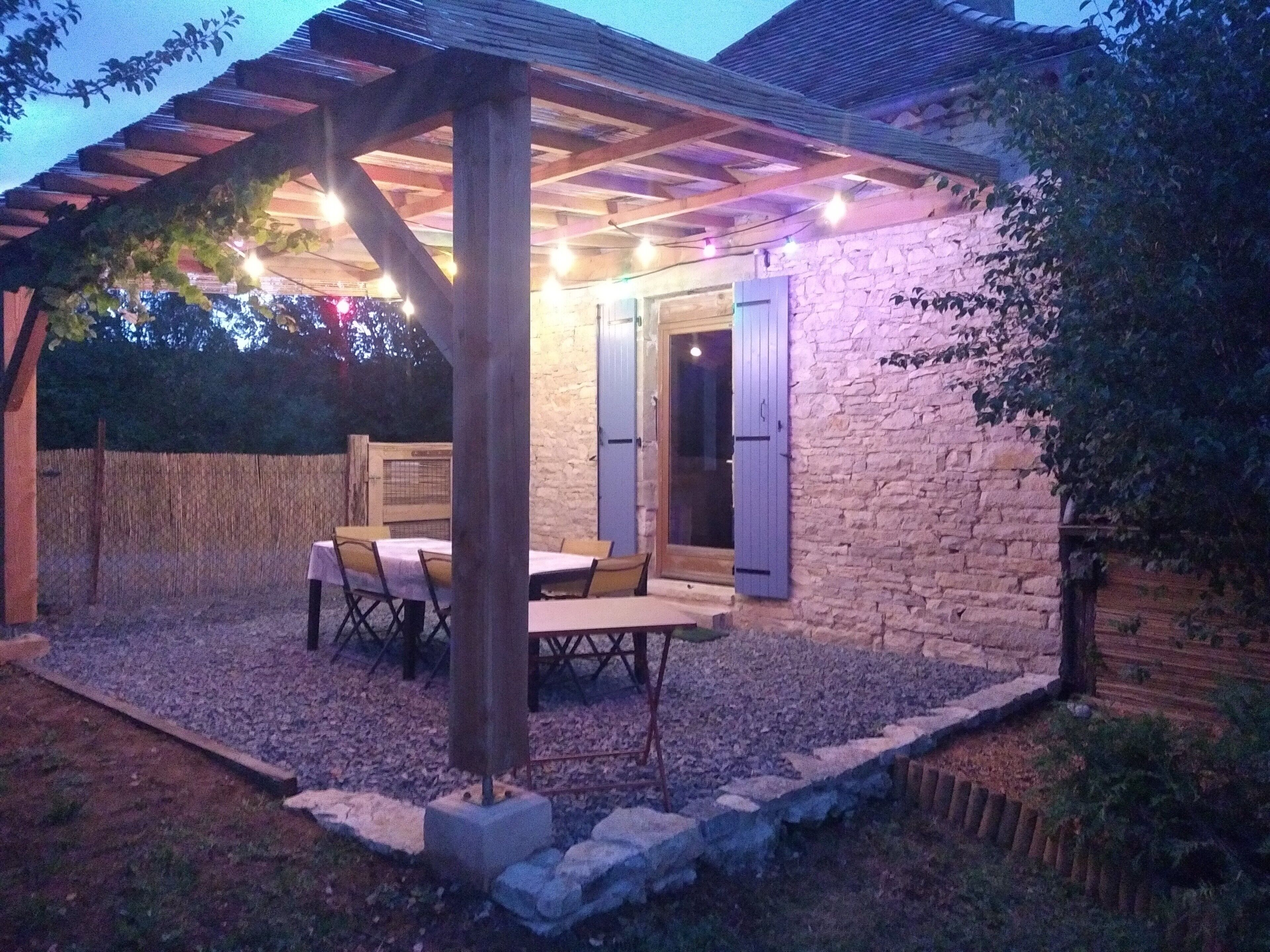Een overdekte buitenruimte om te eten, met een stenen muur, een pergola met lichtjes en een tafel met stoelen.