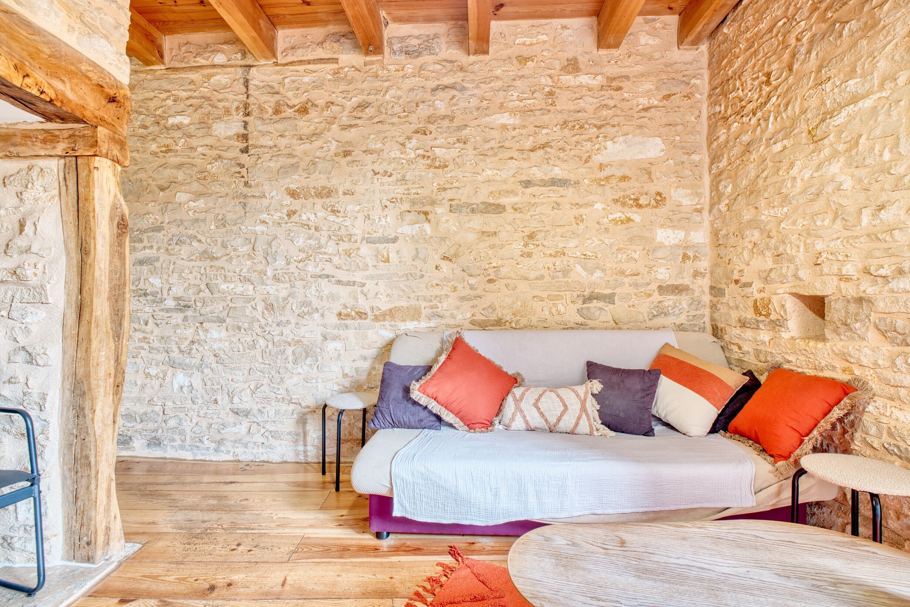 A stone-walled room with a wooden ceiling, a bed with colorful pillows, a small round table, and a chair.
