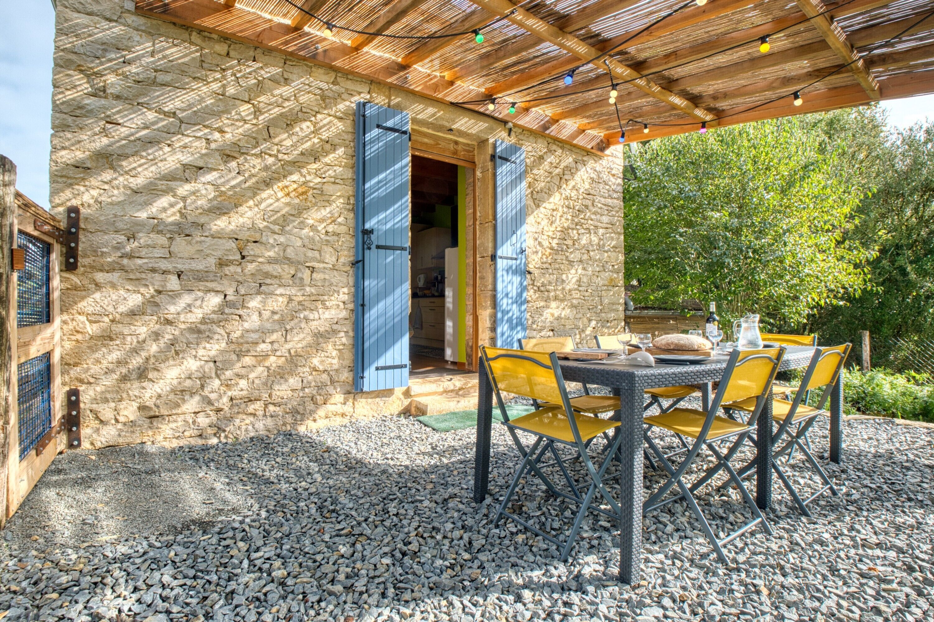 Ein Steinhaus mit einer Holzpergola und einem Tisch mit Stühlen vor der Fassade.