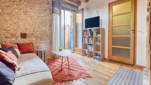A cozy living room with a stone wall, wooden beams, a sofa with cushions, a small table, a bookshelf, and a sliding glass door. - Les Jardins de Boscornut – Organic Farm Cottage near Hautefort in Périgord Noir (Sainte-Orse)
