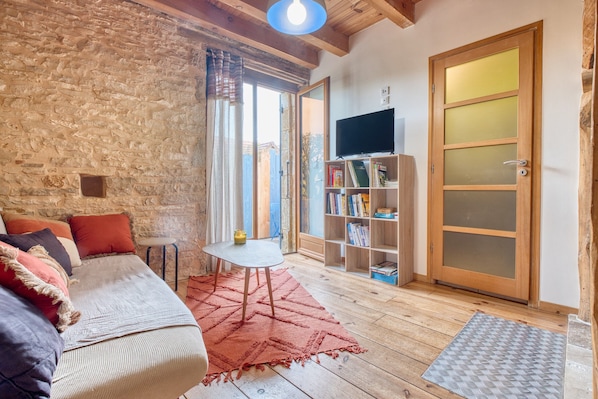 A cozy living room with a stone wall, wooden beams, a sofa with cushions, a small table, a bookshelf, and a sliding glass door.
