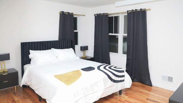 A neatly made bed with white bedding and a gold accent, a bedside table with a lamp, and a window with dark curtains. - Upscale 3-bedroom/2.5-bath unit near EWR, NYC, MetLife, Prudential, and more. (Newark)