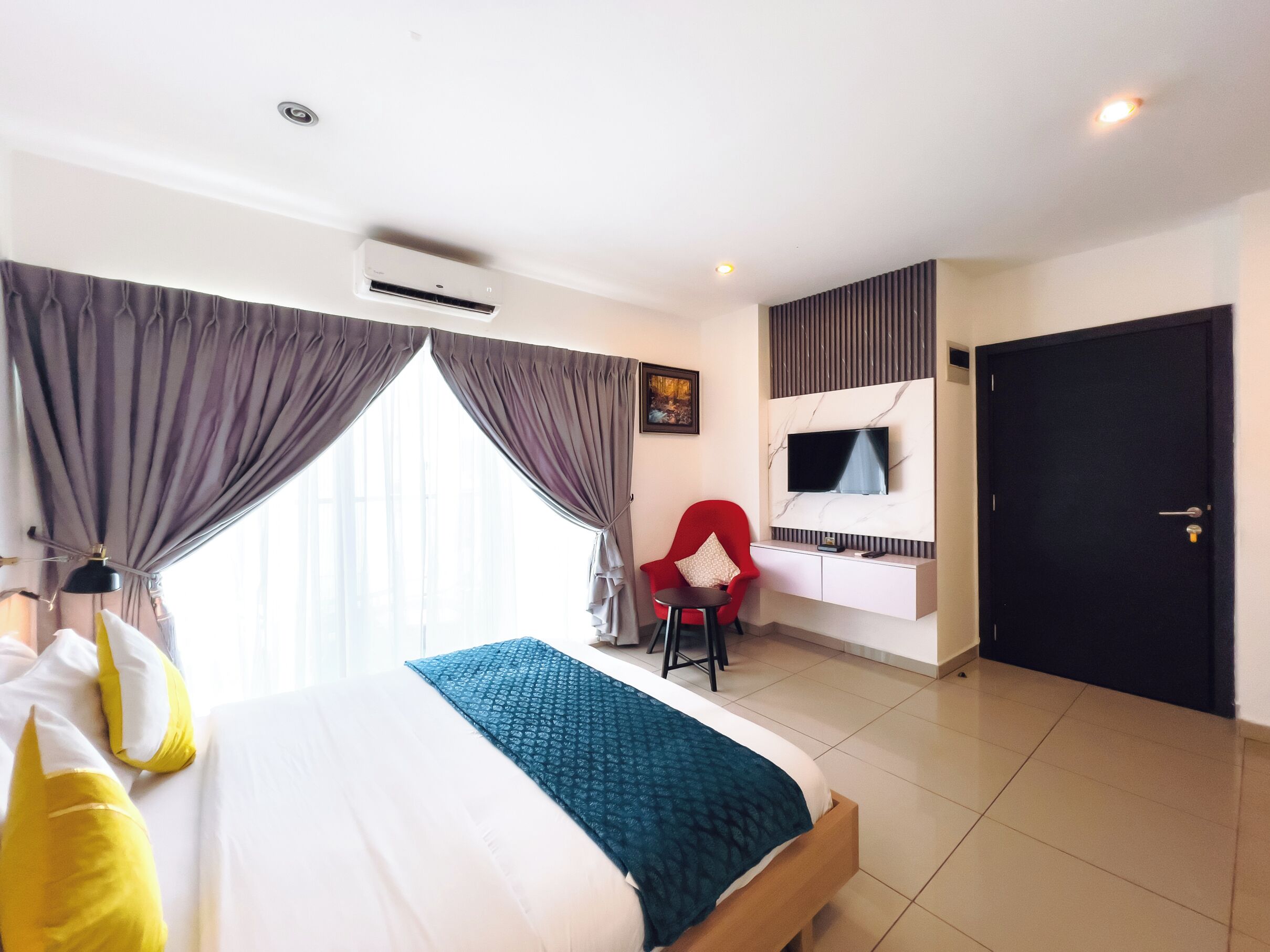 A hotel room with a bed, a red chair, a TV mounted on the wall, and a door.