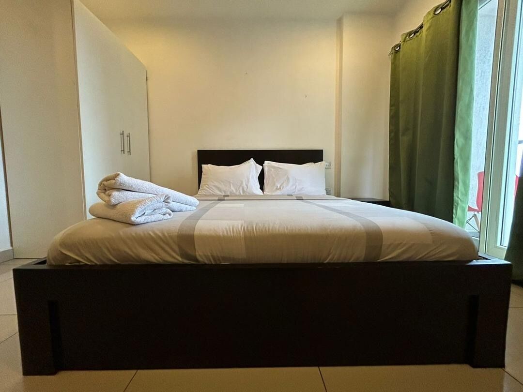 A neatly made bed with white linens and a dark headboard in a room with a green curtain and a window.