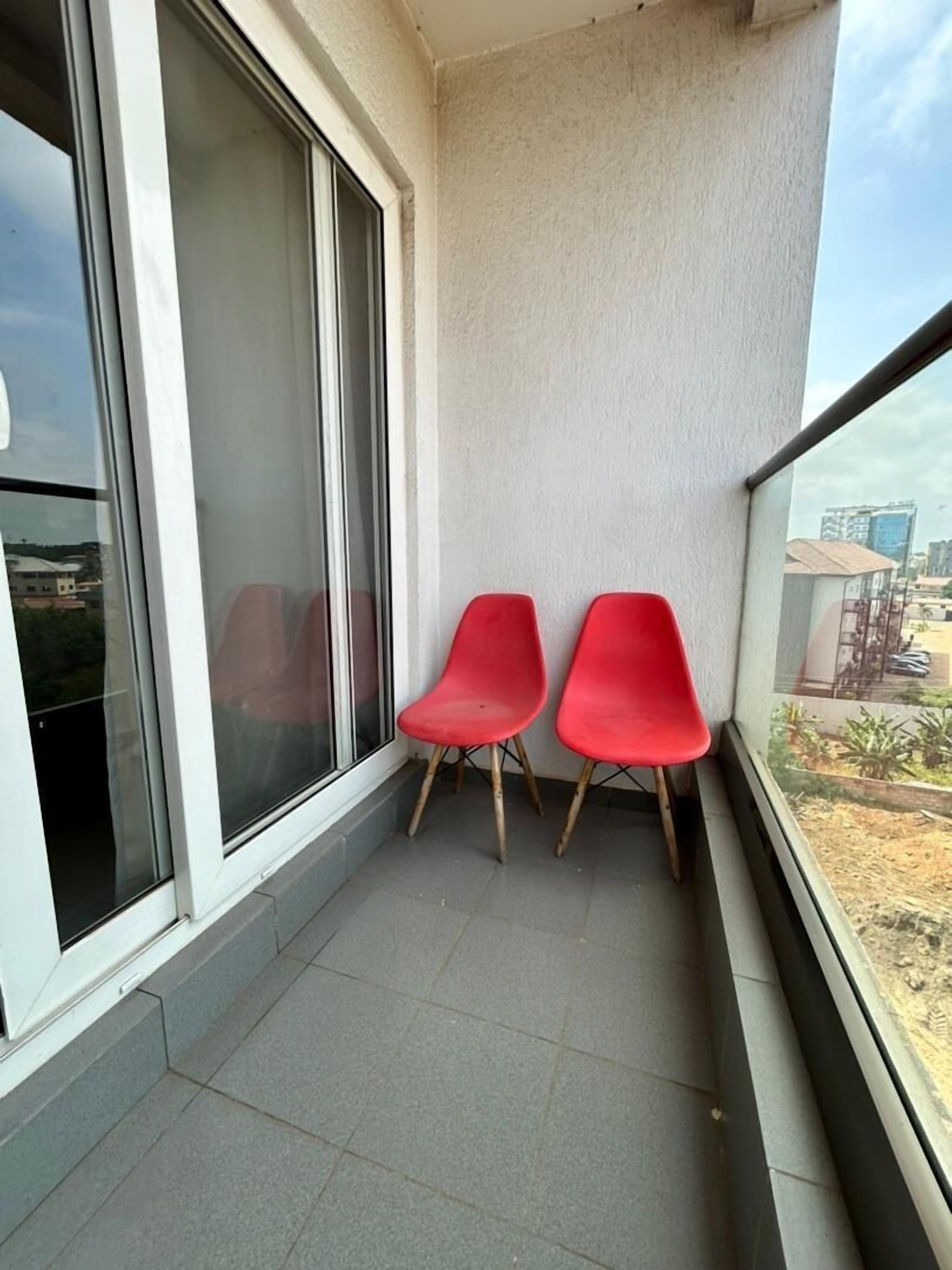 A balcony with two red chairs, a sliding glass door, and a view of buildings and greenery.
