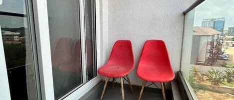 Un balcón con dos sillas rojas, un ventanal corredero y vistas a edificios y vegetación.