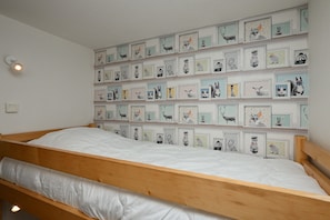 Un letto singolo con struttura in legno e biancheria bianca, una mensola a parete con illustrazioni di animali incorniciate e un piccolo fiorile a parete.