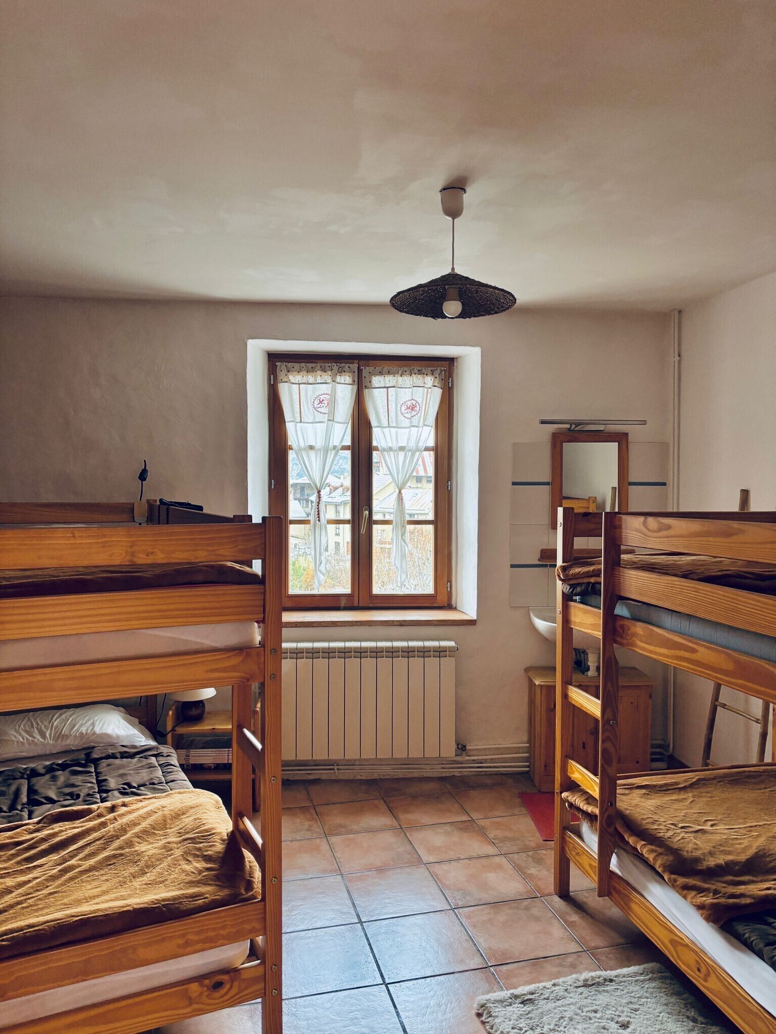 Une chambre de dortoir avec des lits superposés, une fenêtre avec des rideaux, un radiateur, un bureau et un luminaire suspendu.