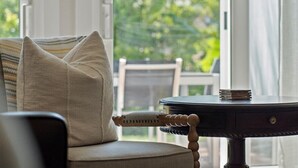 A cozy room with a cushioned chair, a round table with a stack of books, and a view of greenery outside.