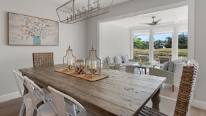 A dining area with a wooden table, white chairs, and a chandelier. There is a painting on the wall and a view of the outdoors through large windows.
