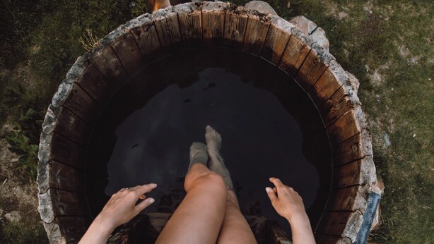Eine Person sitzt in einer runden Holzwanne, die mit Wasser gefüllt ist und von Gras umgeben wird.