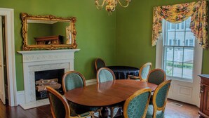 A dining room with a round wooden table, six chairs, a fireplace, a large mirror, and a window with floral curtains. - The Rose Room - Charming Private Studio w/ Ensuite Bath in Historic Home (Clinton)