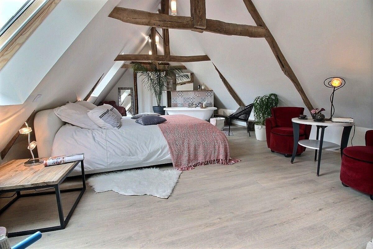 Ein gemütliches, dreieckiges Schlafzimmer im Dachgeschoss mit einem Bett, einem roten Sessel, einem kleinen Tisch und einer weißen Badewanne.