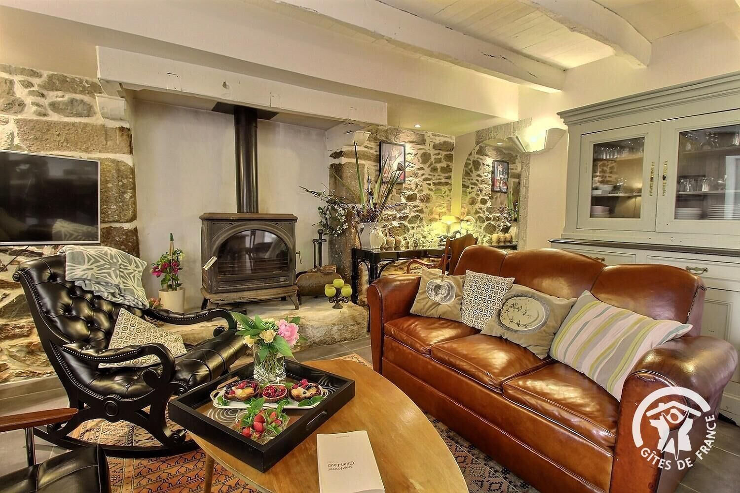 Un salon chaleureux avec un mur en pierre, une cheminée, un canapé en cuir et une table à manger sur laquelle se trouve un plateau de nourriture.
