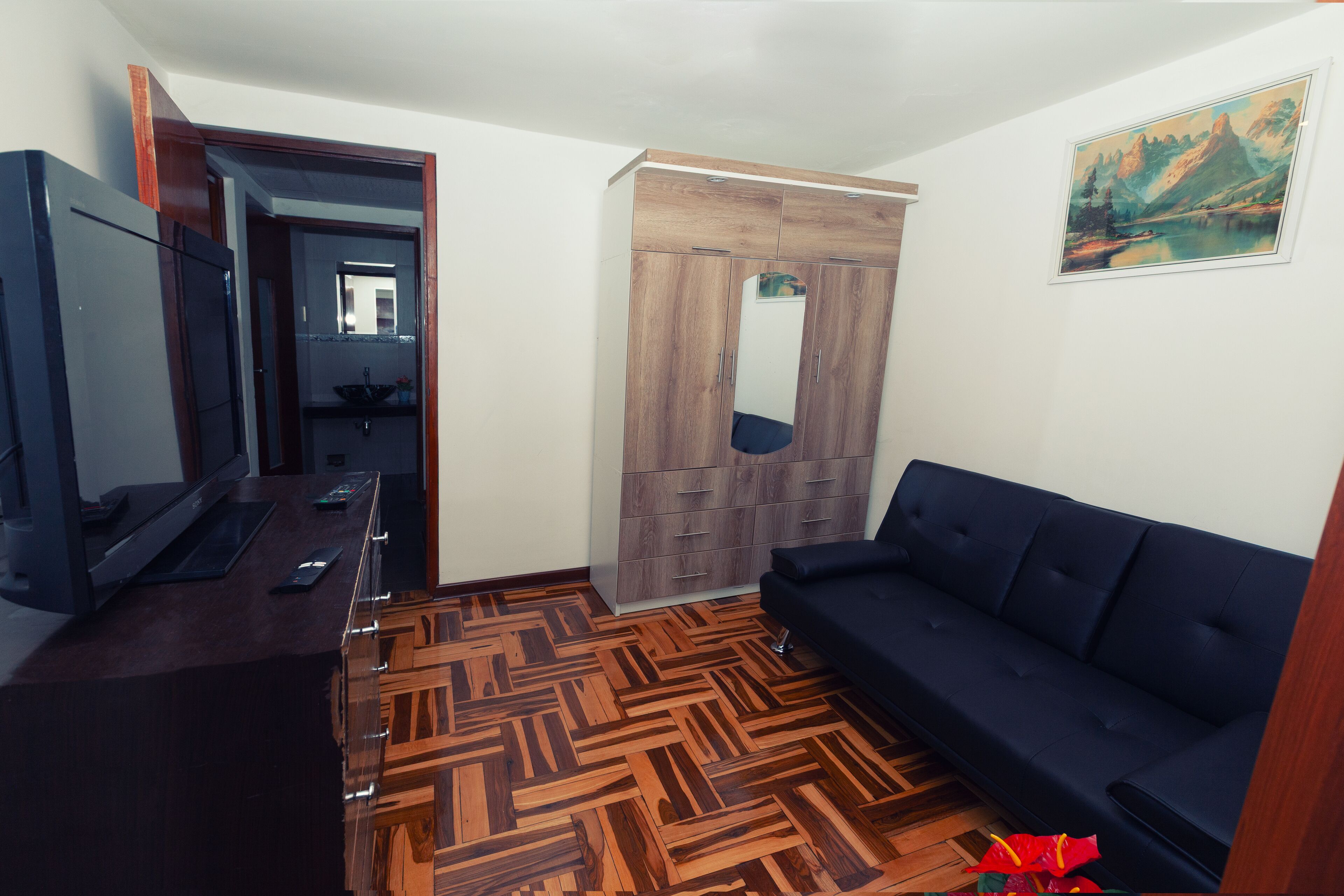 Quarto com piso de madeira em padrão herringbone, sofá escuro, guarda-roupa de madeira, área de cozinha com pia e um quadro na parede.