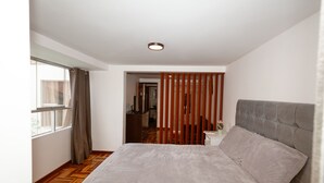 A bedroom with a large bed, a wooden floor, a window with curtains, and a view into another room.