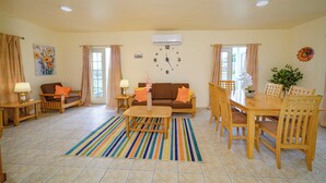 A spacious living room with a sofa, coffee table, dining table, and chairs. A clock is mounted on the wall, and there is a window with curtains. - Charming 2-bedroom apartment with AC, WiFi in fabulous Saint John's (Saint John's)