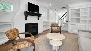 Ein Wohnzimmer mit Kamin, einem darüber montierten Fernseher, Korbsesseln, einem runden Couchtisch und einer Treppe im Hintergrund.
