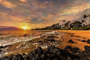 Una playa con costa rocosa, palmeras y un atardecer.