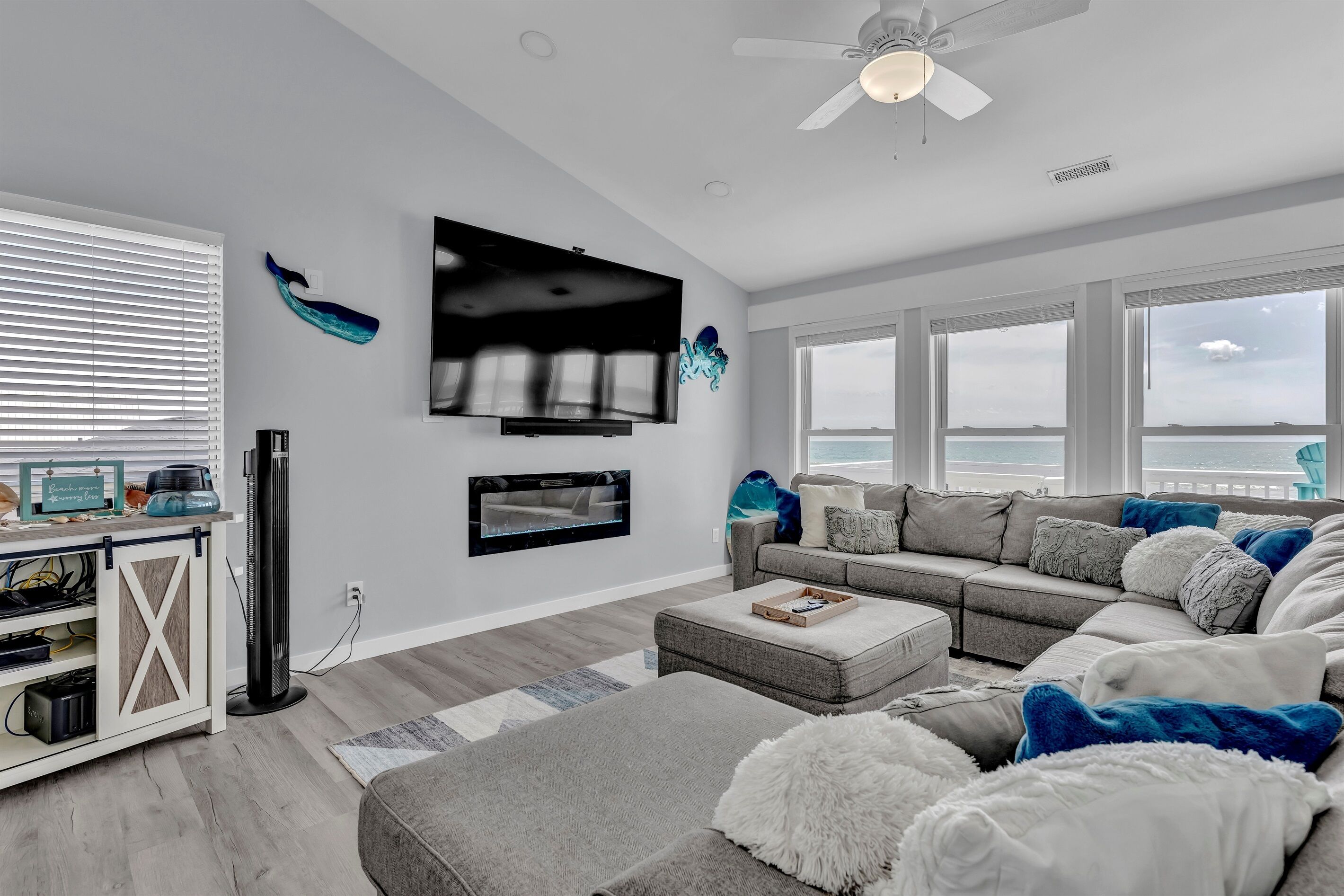 A modern living room with a large flat-screen TV, a ceiling fan, and a view of the ocean through large windows.