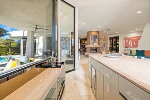 Une cuisine moderne dotée d’un grand îlot central, de portes vitrées donnant sur un coin repas et offrant une vue sur une piscine.