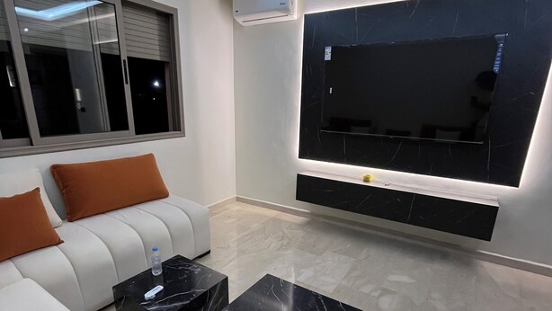Un salon moderne avec un canapé blanc, des tables basses en marbre noir et un grand téléviseur à écran plat fixé au mur.