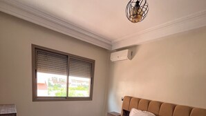 Une chambre à coucher avec un lit, une fenêtre avec des stores, un appareil de climatisation et un luminaire suspendu.