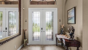 A foyer with a double door, a mirror, a chandelier, a table with decorative items, and a painting on the wall. - Heated Pool, Game Room, & Dock on Freshwater Canal - Mermaid Cove - Roelens (Cape Coral)
