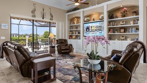 A spacious living room with leather recliners, a glass coffee table, and a large bookshelf filled with decorative items. - Heated Pool, Game Room, & Dock on Freshwater Canal - Mermaid Cove - Roelens (Cape Coral)