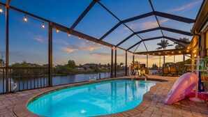 A covered outdoor swimming pool with a view of a lake and palm trees, surrounded by brick flooring and string lights. - Heated Pool, Game Room, & Dock on Freshwater Canal - Mermaid Cove - Roelens (Cape Coral)
