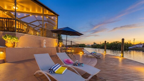 A wooden deck with lounge chairs and a table, overlooking a body of water. - Heated Pool, Game Room, & Dock on Freshwater Canal - Mermaid Cove - Roelens (Cape Coral)