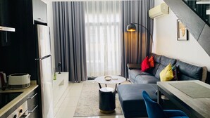 Una sala de estar moderna con sofá, reposapiés y mesa de centro. Cuenta con una zona de cocina equipada con refrigerador y estufa. La habitación tiene ventanas amplias con cortinas.