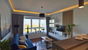 Una sala de estar moderna con un sofá azul, una mesa de centro, un televisor montado en la pared y un ventanal con vista a la ciudad.