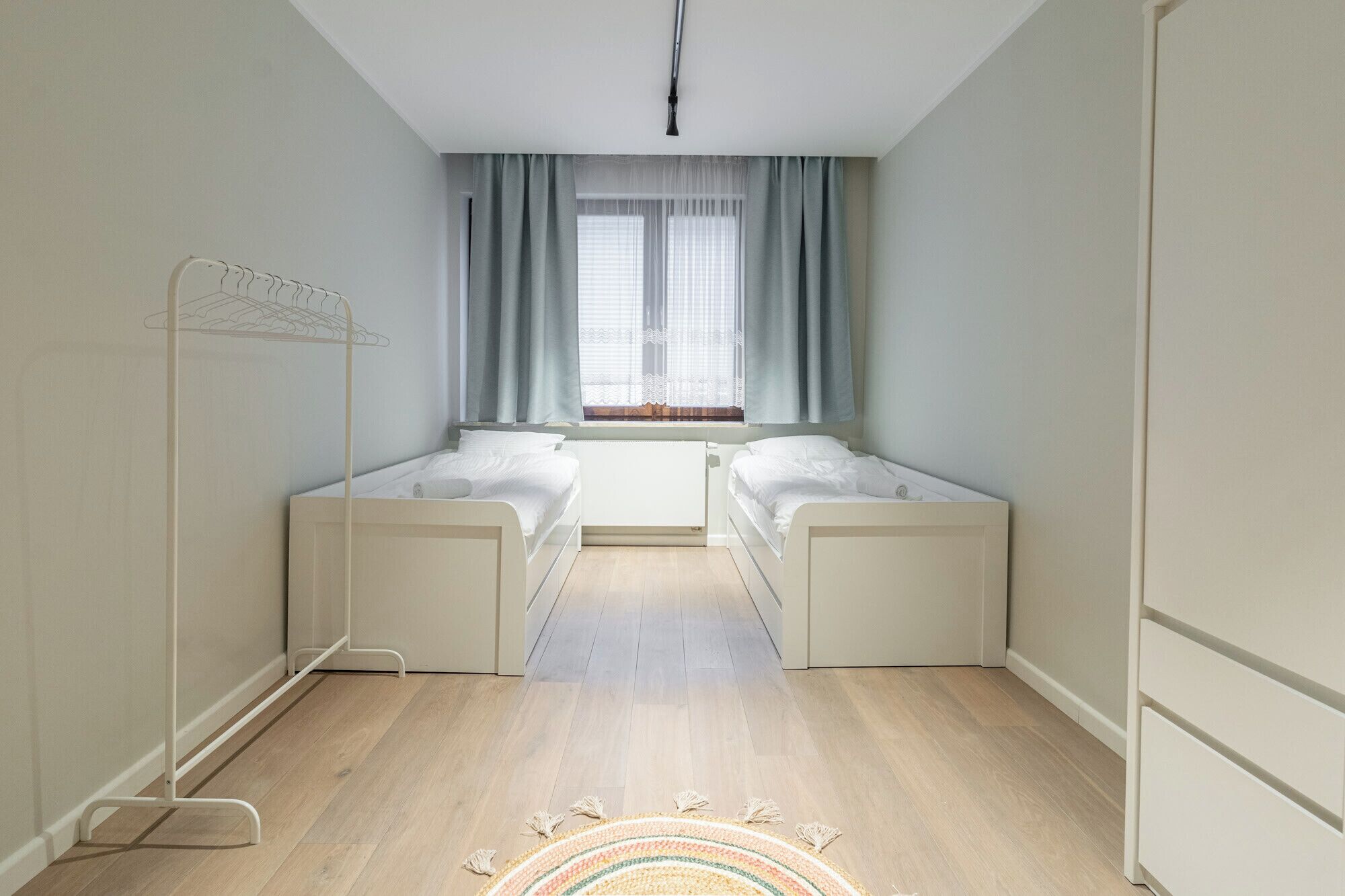A modern, well-lit room with two single beds, a radiator, a window with curtains, and a hanging light fixture.