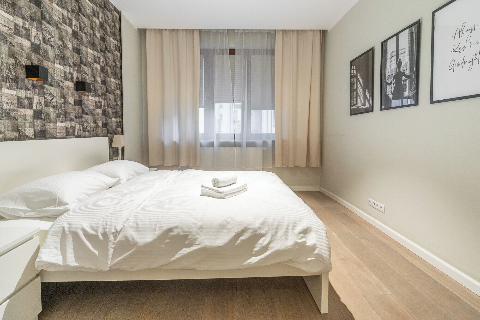 A bedroom with a bed, a nightstand, a wall with a patterned wallpaper, a window with sheer curtains, and framed pictures on the wall.