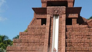 Tobogán de agua con una persona deslizándose, frente a una estructura de piedra con decoración en la parte superior.