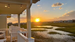 Una terraza de madera con barandillas blancas y luces colgantes, con vista a un cuerpo de agua y casas al fondo durante el atardecer.