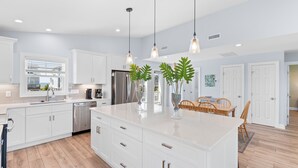 Una cocina moderna con armarios blancos, una isla grande, iluminación colgante y un comedor con sillas de madera.