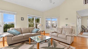 A spacious living room with two beige sofas, a glass coffee table, and a large window offering a view of neighboring houses.