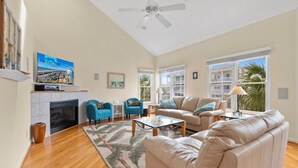 A spacious living room with a fireplace, a ceiling fan, and large windows offering a view of the outside.
